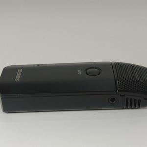 May include: A black Shure brand microphone with a rectangular body, a circular push button on the top surface, and a perforated metal grille at the end. The device features a small circular input port on the side and a smooth matte finish.