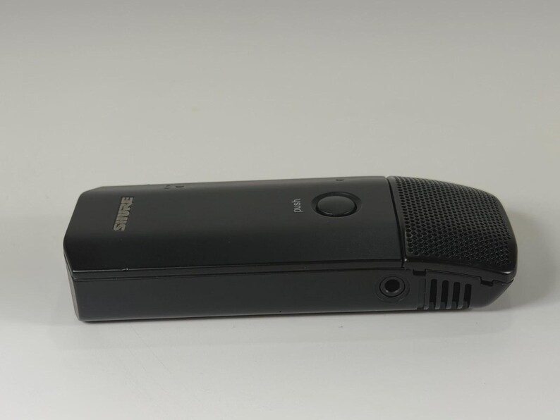 May include: A black Shure brand microphone with a rectangular body, a circular push button on the top surface, and a perforated metal grille at the end. The device features a small circular input port on the side and a smooth matte finish.