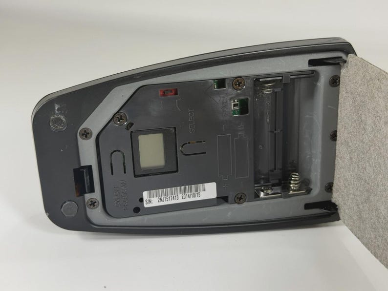 May include: A black electronic device with an open battery compartment, a small square digital display, and a white label featuring the serial number 2NJ1517413 2014/10/15. The internal grey plastic housing includes metal springs and screws.