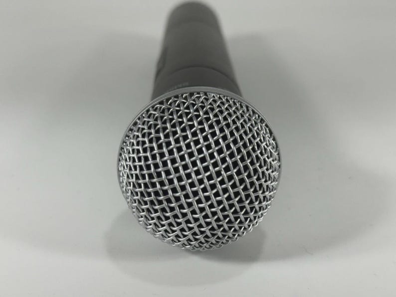 Puede incluir: Primer plano de un micr&oacute;fono gris oscuro. El micr&oacute;fono tiene una cabeza de malla plateada y un cuerpo cil&iacute;ndrico. La cabeza de malla est&aacute; hecha de un metal fino e entrelazado. El micr&oacute;fono est&aacute; dise&ntilde;ado para grabaci&oacute;n vocal o instrumental.