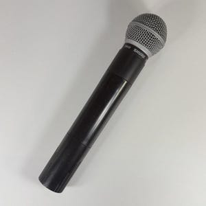 以下が含まれることがあります： 銀色の球状ワイヤーメッシュグリルを備えた黒いハンドヘルドダイナミックマイク。マイク本体の上部、グリルのすぐ下にSHURE SM58という白い文字が印刷されており、モデル名とブランド名を示しています。