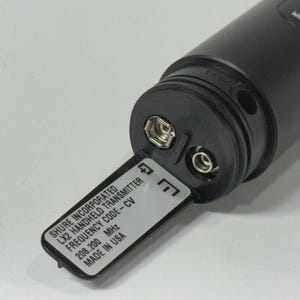 May include: A black Shure LX2 handheld transmitter with an open battery compartment. The label reads "SHURE INCORPORATED LX2 HANDHELD TRANSMITTER FREQUENCY CODE-CV 208.200 MHz MADE IN USA". The device has a mute button.