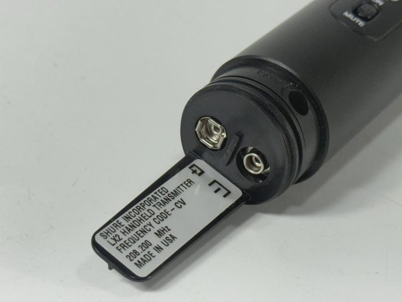 May include: A black Shure LX2 handheld transmitter with an open battery compartment. The label reads "SHURE INCORPORATED LX2 HANDHELD TRANSMITTER FREQUENCY CODE-CV 208.200 MHz MADE IN USA". The device has a mute button.
