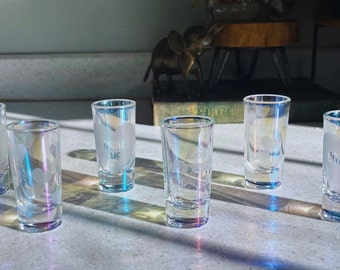 May include: Six clear glass shot glasses with frosted white hearts and text. The glasses are arranged in a row on a white surface. The text on the glasses reads "Party Hardy", "Drink Me", "Ditties Up", and other phrases.