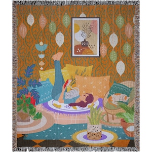 May include: A woven tapestry throw blanket featuring a colorful illustration of a woman relaxing on a couch with a cat, surrounded by plants and pillows. The background is a patterned orange wall with hanging leaves.
