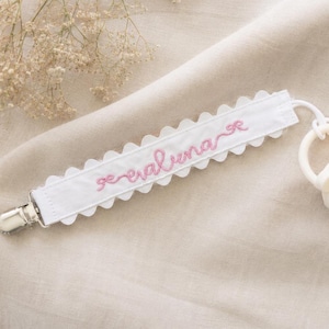 May include: A white fabric pacifier clip featuring a scalloped edge and the name "evaluna" embroidered in pink thread. The strap is attached to a silver metal clip on one end and a cream-colored silicone pacifier on the other end.