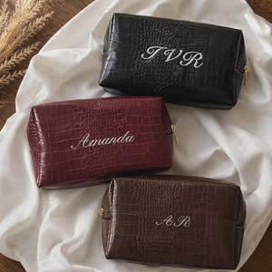 May include: Three rectangular cosmetic bags with a crocodile-embossed texture. One bag is black with the embroidered text TVR, one is burgundy with the embroidered name Amanda, and one is dark brown with the embroidered initials AR. Each has a gold zipper.