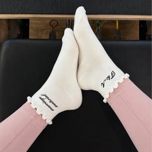 May include: A pair of off-white ankle socks with ruffled cuffs. The left sock features the black text "marriage material" and the right sock features the black text "T&A" in a cursive font. The socks are worn with light pink leggings.
