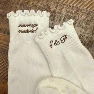 May include: A pair of white ankle socks with ruffled cuffs. One sock features the brown embroidered text "marriage material" and the other features the brown embroidered text "G & F". The soles have small, circular, light-colored grip dots.