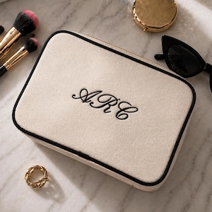 May include: A rectangular off-white canvas travel case with black piping and a gold-tone zipper. The center of the case features the embroidered black script letters ARC. The case has a structured design with rounded corners and a flat top surface.