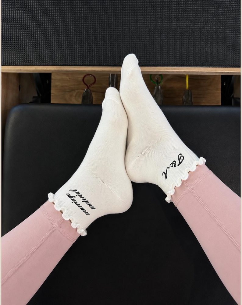 May include: A pair of off-white ankle socks with ruffled cuffs. The left sock features the black text "marriage material" and the right sock features the black text "T&A" in a cursive font. The socks are worn with light pink leggings.