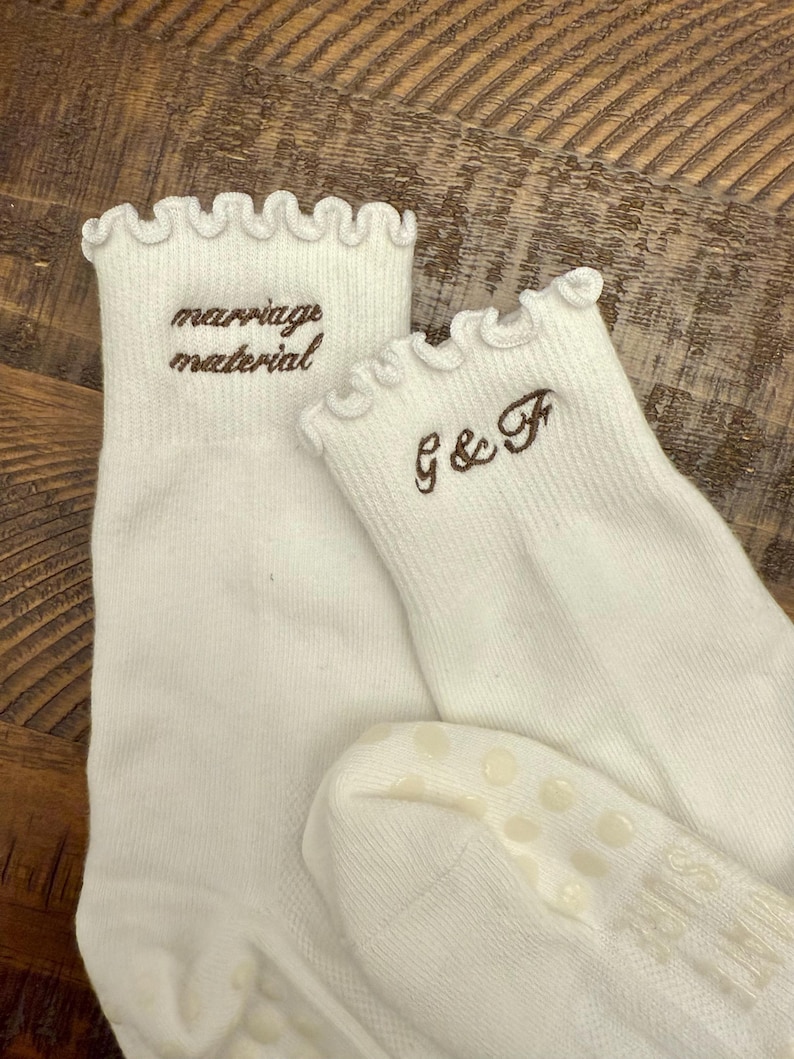 May include: A pair of white ankle socks with ruffled cuffs. One sock features the brown embroidered text "marriage material" and the other features the brown embroidered text "G & F". The soles have small, circular, light-colored grip dots.