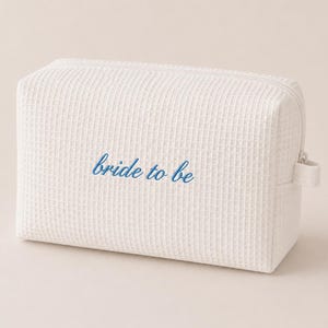 May include: A white, rectangular cosmetic bag with a textured waffle weave pattern. The words "bride to be" are embroidered in blue on the front. The bag has a zipper closure and a small fabric loop on the side.