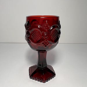 Puede incluir: Una copa de color rojo rubí con detalles ornamentados y texturizados. El vidrio presenta una base hexagonal y un tallo que conduce a un cuenco detallado. La copa es probablemente de vidrio prensado y es adecuada para servir bebidas.