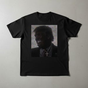 May include: Black t-shirt featuring a portrait of a smiling person with a cross pendant. The portrait is in a rectangular frame. The image has a grainy, slightly distorted effect.