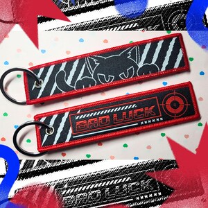 May include: Two rectangular embroidered keychains with red borders and silver metal rings. One features a black cat graphic with white claw marks. The other displays the text "BAD LUCK" in red letters alongside a red circular target symbol.