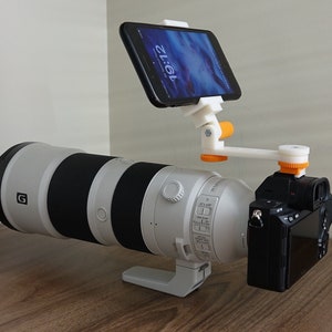 Könnte beinhalten: Ein weiß-schwarzes Kameraobjektiv, das an einer schwarzen Kamera befestigt ist, mit einem weißen und orangefarbenen Telefonhalter, der oben auf der Kamera befestigt ist. Der Telefonhalter hält ein schwarzes Smartphone mit einem weißen Bildschirm, auf dem die Zeit 19:12 angezeigt wird.