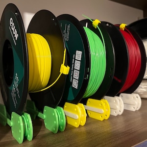 Puede incluir: Un conjunto de cuatro soportes para carretes de filamento para impresora 3D. Los soportes están hechos de plástico verde y tienen una ranura para que el carrete se asiente. Los carretes son negros con filamento amarillo, verde y rojo.