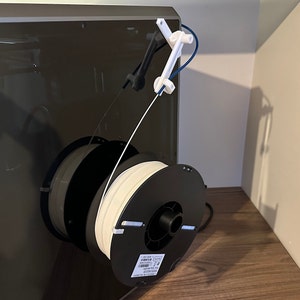 Könnte beinhalten: Eine weiße Filamentspule ist auf einem schwarzen Spulenhalter montiert, der an einem 3D-Drucker befestigt ist. Das Filament wird durch einen weißen und schwarzen Filamentführer geführt.