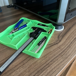 Puede incluir: Un organizador de herramientas de plástico verde con compartimentos para varias herramientas, incluyendo un calibre digital, destornilladores y un par de alicates. El organizador está sentado sobre una superficie de madera.