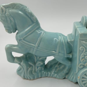Könnte beinhalten: Hellblauer Keramik-Pflanztopf in Form eines Pferdes, das einen Wagen zieht. Das Pferd bäumt sich auf, und der Wagen hat dekorative Reliefschnitzereien von Engeln und Musikinstrumenten. Der Pflanztopf hat vier Räder.