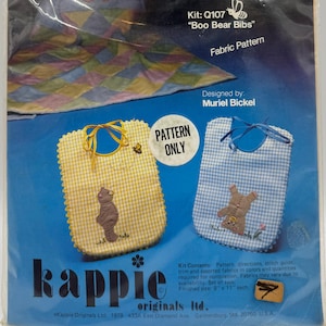 May include: A sealed package of a fabric pattern for making baby bibs. The package features two bib designs: one yellow gingham and one blue gingham, each with a teddy bear applique. The text "Boo Bear Bibs" is visible.