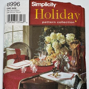 Op de afbeelding: Een Simplicity Holiday patrooncollectie omslag. De afbeelding toont een feestelijke tafelsetting met een rode tafelloper, decoratieve kussens en een bloemstuk. De tekst "Holiday pattern collection" is in goud weergegeven.