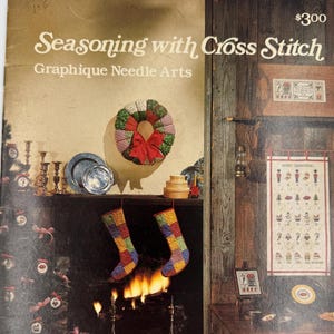 May include: The cover of a cross-stitch book titled "Seasoning with Cross Stitch" by Graphique Needle Arts. The cover features a cozy fireplace scene with stockings, a wreath, and a Christmas tree. The price is $300.