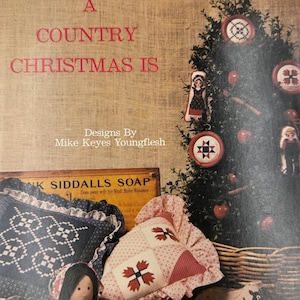 Op de afbeelding: Een vintage boekomslag met de titel "A Country Christmas Is" en de naam van de auteur. De afbeelding toont een kerstboom met ornamenten, kussens, een pop en een houten speelgoed. De omslag staat op een jute achtergrond.
