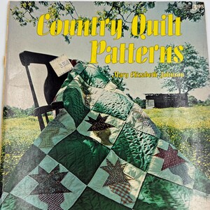 May include: A vintage magazine titled "Country Quilt Patterns" featuring a green and patterned quilt draped over a chair. The cover has yellow text and a pastoral scene background. The quilt has star-shaped patterns.