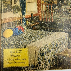 Puede incluir: Una portada de libro vintage titulada "Tell City Primer of Early American Home Decorating". La portada presenta una ilustración de un dormitorio con ropa de cama floral, cortinas azules y una pequeña mesita de noche de madera. La paleta de colores incluye beige, azul y amarillo.