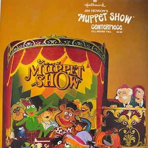 Könnte beinhalten: Ein Hallmark Jim Henson's Muppet Show Tischaufsteller mit einer Höhe von 31,75 cm. Das Pappdisplay zeigt eine farbenfrohe Illustration von Muppet-Figuren, die auf einer Bühne mit roten Vorhängen und einem Balkon mit zwei älteren Figuren auftreten.