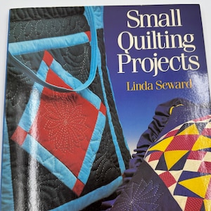 May include: A book titled "Small Quilting Projects" by Linda Seward. The cover features images of quilted items, including a tote bag with red and blue accents, and a pillow with yellow, red, and blue geometric patterns. The background is a light beige.