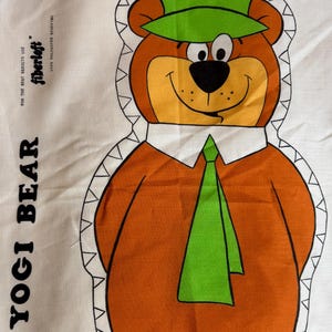 Puede incluir: Panel de tela con una imagen de dibujos animados de Yogi Bear. El oso está representado en tonos naranja, marrón y verde, con un sombrero y una corbata verdes. El texto "YOGI BEAR" está impreso a la izquierda, con instrucciones "DEJAR ABIERTO PARA RELLENAR" en la parte inferior.