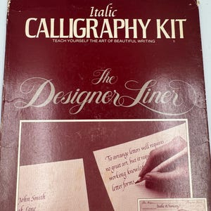 Puede incluir: Un kit de caligrafía vintage titulado "Italic Calligraphy Kit" con el texto "Teach Yourself the Art of Beautiful Writing". El kit incluye un "Designer Liner" y hojas de práctica. La portada es de color rojo oscuro con texto e ilustraciones en blanco y crema.