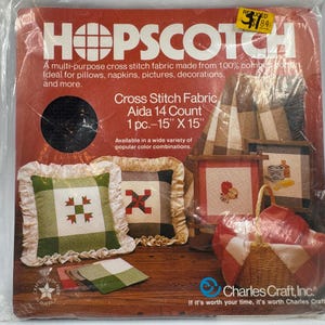 May include: A package of Hopscotch cross stitch fabric. The red package features the brand name in white block letters. The package includes a 15" x 15" piece of Aida 14 count fabric. The package also shows examples of finished cross-stitch projects.