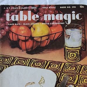 Puede incluir: Un folleto de patrones de ganchillo vintage titulado "Table Magic" con un mantel individual de ganchillo marrón y amarillo, un vaso de agua con un posavasos de ganchillo y un cuenco de fruta.