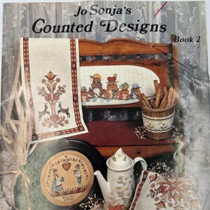 May include: A softcover craft book titled Jo Sonja's Counted Designs Book 2. The cover features a collection of cross-stitch projects including a table runner, a round wall hanging, a small wooden plaque, and a teapot with a matching teacup.