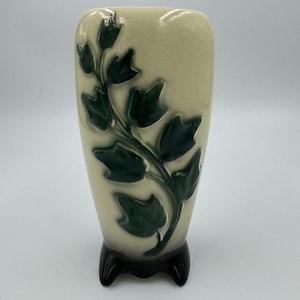 Może przedstawiać: Kremowy ceramiczny wazon z ciemnozielonym wzorem liści. Wazon ma prostokątny kształt z zaokrąglonymi krawędziami i stoi na czarnej, trójnożnej podstawie. Liście są szczegółowe i dodają projektowi odrobinę natury.