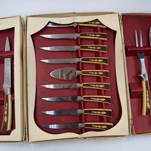 May include: A vintage cutlery set in a cream-colored box with a red interior. The set includes various knives, a cake server, and a carving fork. The handles are a combination of brown and cream with a decorative gold leaf design.