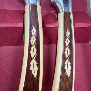 May include: Two knives with stainless steel blades and cream-colored handles. The handles have a dark wood inlay with a leaf design. The knives are set against a red background.