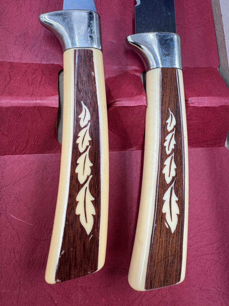 May include: Two knives with stainless steel blades and cream-colored handles. The handles have a dark wood inlay with a leaf design. The knives are set against a red background.