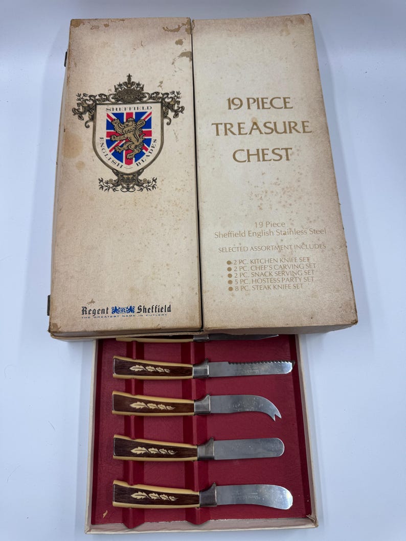 May include: A vintage 19-piece Sheffield English Stainless Steel Treasure Chest set. The box is beige with text and a crest. Inside, a red tray holds knives with brown and cream handles.