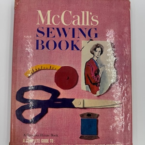 Op de afbeelding: Een vintage McCall's Sewing Book met een roze omslag. De omslag bevat illustraties van naai-instrumenten zoals een schaar, een meetlint en een klos garen. De titel van het boek staat in grote witte letters. Het boek is een complete gids voor kleding maken, kleermaken, repareren, borduren en interieurdecoratie.