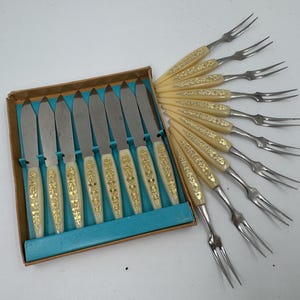 Op de afbeelding: Een vintage bestekset met botermessen en vorken. De messen zitten in een doos met crèmekleurige handvatten en gouden bloemmotieven. De vorken hebben bijpassende handvatten. De set is op een wit oppervlak geplaatst.