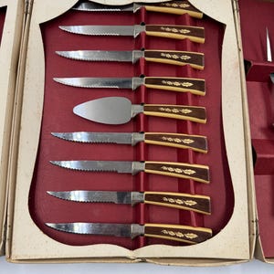 May include: A vintage cutlery set in a cream-colored box with a red interior. The set includes serrated knives and a cake server. The handles are a combination of cream and brown with a leaf design. The box is open, revealing the arrangement of the knives.