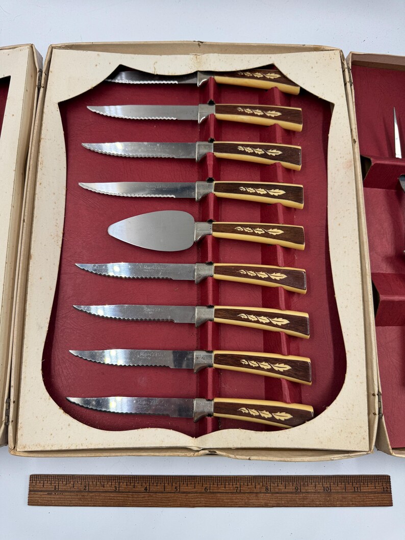 May include: A vintage cutlery set in a cream-colored box with a red interior. The set includes serrated knives and a cake server. The handles are a combination of cream and brown with a leaf design. The box is open, revealing the arrangement of the knives.