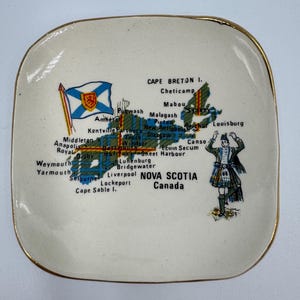 May include: A square, off-white ceramic plate with a gold rim. The plate features a map of Nova Scotia, Canada, with city names and a flag. A figure in traditional Scottish attire is depicted on the right side of the plate.
