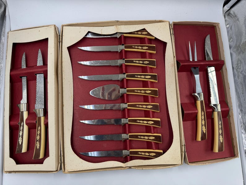 May include: A vintage cutlery set in a cream-colored box with a red interior. The set includes various knives, a cake server, and a carving fork. The handles are a combination of brown and cream with a decorative gold leaf design.