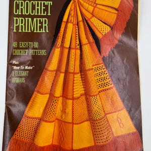 May include: A vintage crochet primer book with a brown cover and the title "CROCHET PRIMER" in green. The book features a folded orange and yellow afghan with fringe. The cover text includes "48 EASY-TO-DO CROCHET PATTERNS" and brand names.